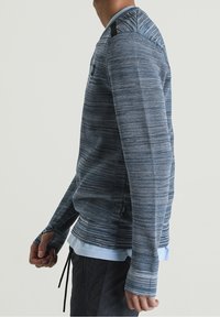 Niebieski sweter dzianinowy z teksturowanym wzorem, z czarnymi akcentami na ramionach i wydłużonymi mankietami, noszony na jasnoniebieskiej koszuli.