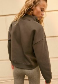 Sweatshirt vert olive avec un col montant, coupe oversize et ourlet côtelé. Porté avec des leggings gris texturés. Tissu doux et design minimaliste.