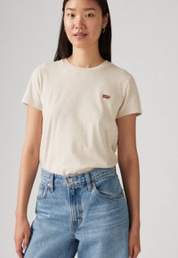 Beige bomulls t-skjorte med rund hals og korte ermer, med et lite rødt Levi's-logo på venstre bryst. Kombinert med lyseblå jeans.