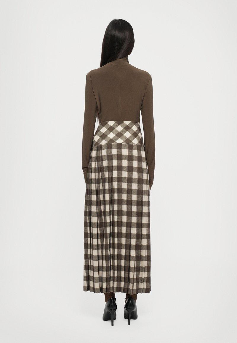 Vestido marrón de manga larga con un top ajustado y una falda de cuadros con tablas en tonos marrón y crema, que presenta un escote alto y zapatos de tacón negros.