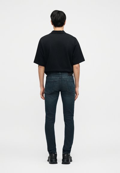 Μαύρο κοντομάνικο πόλο, στενά τζιν από σκούρο τζιν και μαύρα αθλητικά παπούτσια. Το σύνολο χαρακτηρίζεται από απλό σχέδιο με ελάχιστη λεπτομέρεια.