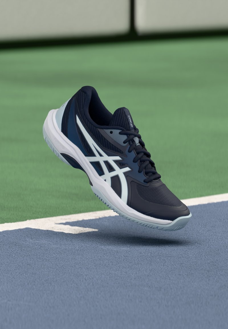 Scarpa sportiva blu navy realizzata in mesh e gomma, con dettaglio grigio, design con lacci e suola con texture, posizionata sopra un campo da tennis.