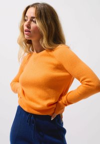 Maglione di lana arancione con collo a giro e maniche lunghe, caratterizzato da una texture morbida e una silhouette aderente. Abbinato a pantaloni blu navy.