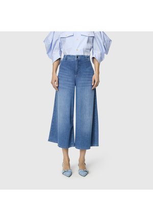 Person trägt weit geschnittene, verkürzte blaue Jeans, ein hellblaues Hemd mit Puffärmeln und spitze blaue Schuhe mit Schnallen.