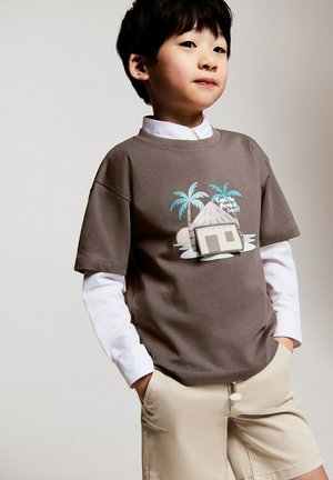 Băiat tânăr purtând un tricou maro cu grafica unei colibe și a unei palme peste un tricou alb cu mâneci lungi, asortat cu pantaloni scurți bej, cu mâinile în buzunare.