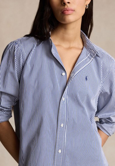 Mladá žena nosí modro-bielu vertikálne pruhovanú košeľu na gombíky s vyhrnutými rukávmi a malým vyšívaným logom na hrudi.