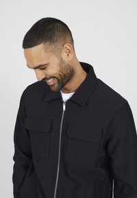 Schwarze gerippte Jacke mit Kragen, zwei Fronttaschen und einem Reißverschluss. Der Stoff hat eine strukturierte Textur, die ein strukturiertes Aussehen erzeugt.