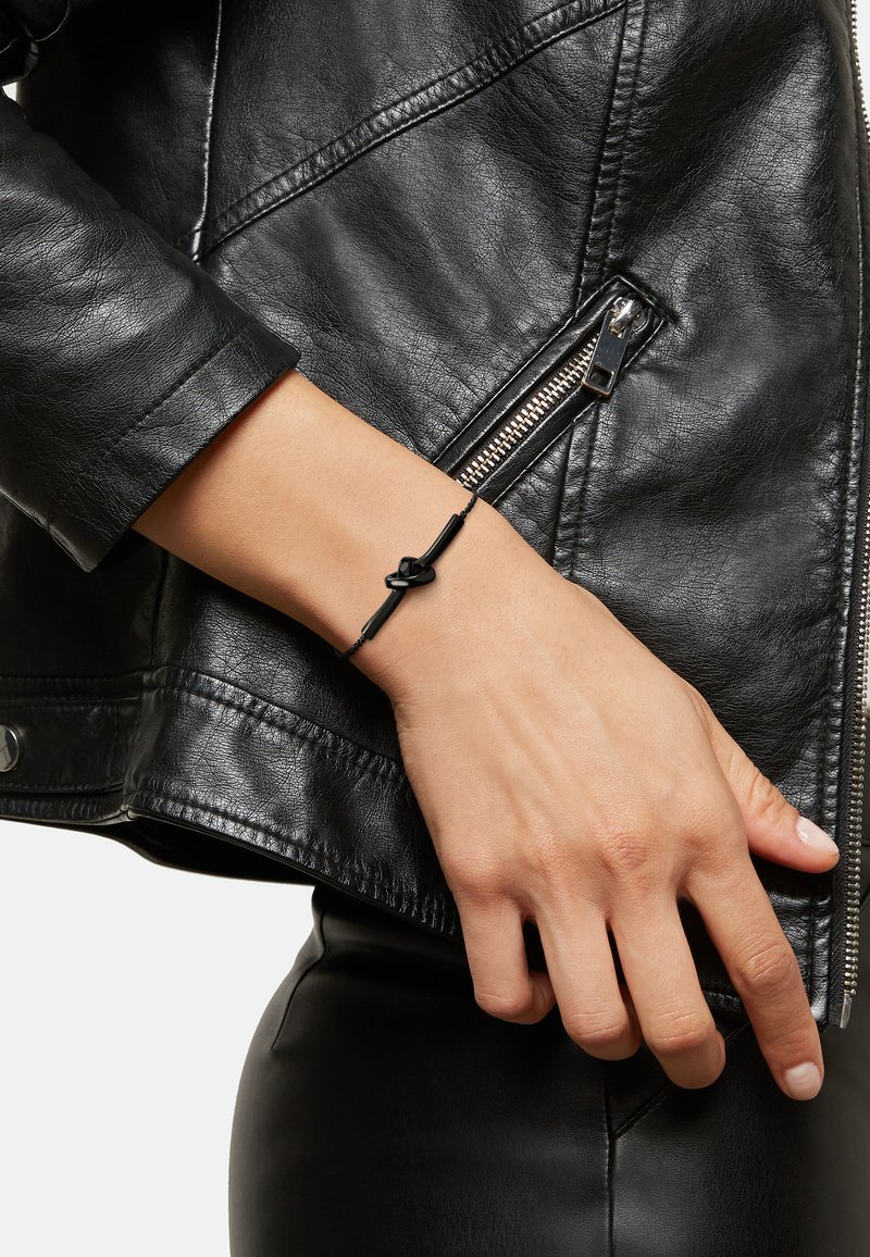 Černý kožený náramek s designem uzlu, nošený na zápěstí, v kombinaci s černou koženou bundou, zdůrazňující elegantní a minimalistický styl.