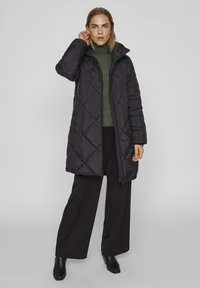 Črn prešiten dolg plašč z visokim ovratnikom, s kritjem z zadrgo in stranskimi žepi, položen preko zelene pletene jopice in črnih hlač.