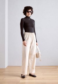 Čierne dlhé rukávy, krémové široké nohavice, čierne ploché topánky a slnečné okuliare, v kombinácii s malou béžovou crossbody taškou. Pozadie z drevenej podlahy.