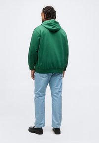 Άνδρας με πλεγμένα μαλλιά που φοράει πράσινο φούτερ με κουκούλα, ανοιχτόχρωμο μπλε τζιν και μαύρα παπούτσια, đứng γυρισμένος προς τα πίσω πάνω σε λευκό φόντο.