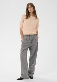 Pfirsichfarbener, gerippter Pullover mit kurzen Ärmeln, kombiniert mit lockeren grauen Hosen. Das Outfit umfasst einen hellen Gürtel und schwarze spitze Schuhe.