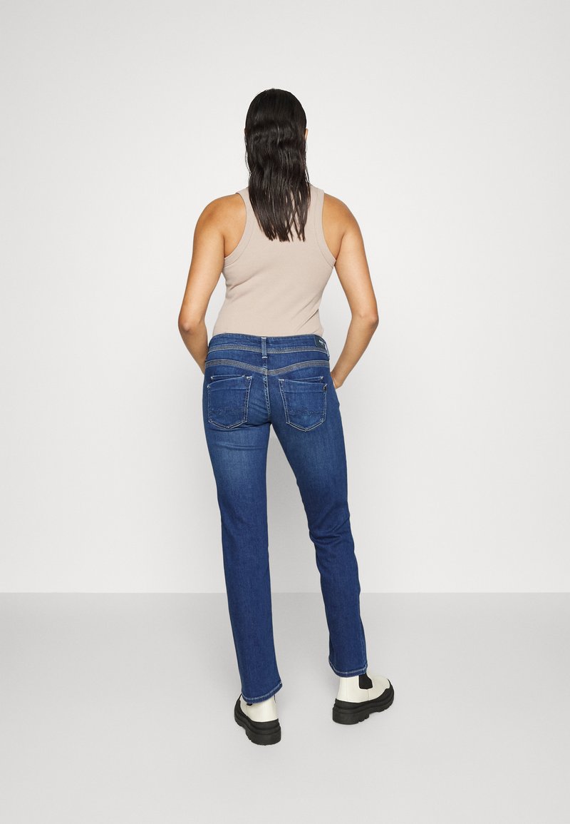Jeans en denim bleu foncé avec une coupe droite, dotés de poches arrière, associés à un débardeur beige et des bottines noires à la cheville.