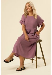 Lungo vestito viola con maniche corte a volant, tessuto strutturato e vita strizzata. Indossato con sandali beige a incrocio. Seduta su uno sgabello di legno.