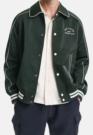 Mandlig torso iført en mørkegrøn varsityjakke med hvide piping og trykknapper over en hvid poloshirt og marineblå cargobukser.
