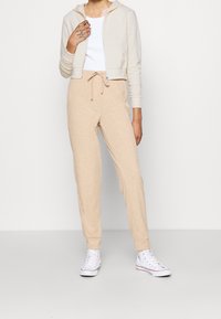 Persona che indossa pantaloni jogger beige a coste, canotta bianca, giacca a zip beige cropped e sneakers alte bianche, in piedi contro un muro bianco semplice.