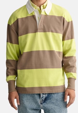 Man in een gele en bruine brede horizontaal gestreepte longsleeve rugbyshirt met witte kraag en lichtblauwe spijkerbroek.