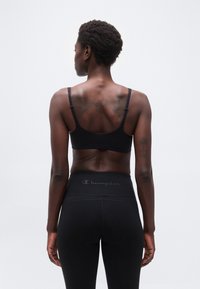 Γυναίκα που στέκεται γυρισμένη προς τα πίσω φορώντας μαύρο αθλητικό μπουστάκι και μαύρο κολάν με ψηλή μέση, με ορατά τατουάζ στην πλάτη και τα χέρια.