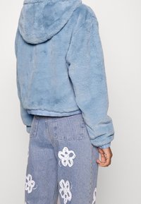 Jasnoniebieska kurtka z sztucznego futra z kapturem, o krótkim kroju i ściągaczami na rękawach, zestawiona z jasnymi jeansami zdobionymi białymi wzorami kwiatowymi.