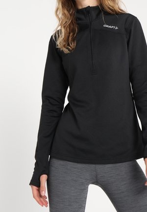 Kobieta w czarnym sportowym topie z długim rękawem, półzamkiem i logo "CRAFT", połączonym z przylegającymi szarymi legginsami.