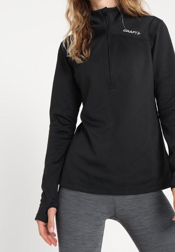 Mujer con una camiseta deportiva negra de manga larga con cremallera hasta la mitad y el logo "CRAFT", combinada con leggings ajustados grises.
