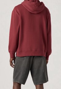Bordeauxrotes Kapuzensweatshirt mit lockerer Passform und gerippten Bündchen, kombiniert mit grauen Jeansshorts, von hinten gesehen. Keine sichtbaren Muster oder Akzente.