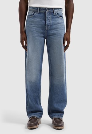 Man in blauwe jeans met wijde pijpen, wit shirt en bruine leren mocassins, staand tegen een lichte achtergrond.