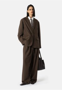 Hnědý oversized oblek s jedním knoflíkem, dvěma předními kapsami a bílou košilí s vzorovanou kravatou. Doplněno o černé boty a tašku.