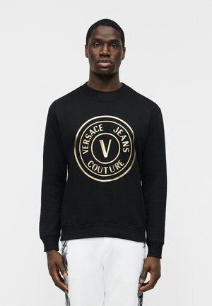 Moški v črni majici s kapuco z zlatim logotipom Versace Jeans Couture na sredini prsnega koša, kombinirana z belimi hlačami s črnimi stranskimi vzorci.