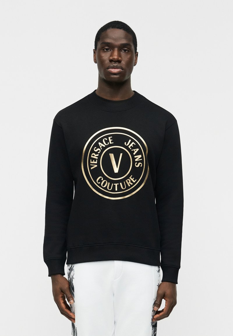 Άνδρας που φοράει μαύρο φούτερ με χρυσό λογότυπο Versace Jeans Couture στο κέντρο του στήθους, συνδυασμένο με λευκό παντελόνι με μαύρα πλευρικά σχέδια.