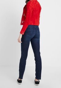 Giacca da motociclista in camoscio rosso con tasche con cerniera, abbinata a jeans skinny blu scuro e sneakers nere. Texture liscia e design aderente.