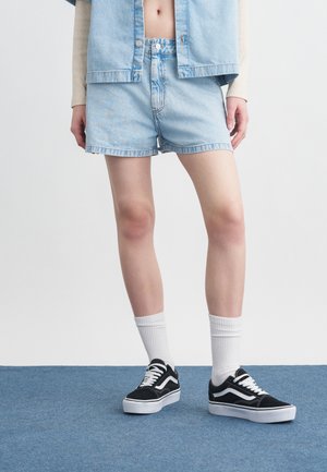 Άτομο που φοράει ανοιχτό μπλε τζιν σορτς και τζάκετ, λευκές κάλτσες και μαύρα αθλητικά παπούτσια χαμηλού προφίλ, στέκεται σε μπλε χαλί.