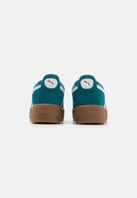 Teal semskinnesko med hvite detaljer og såle av brun gummi, med en sirkulær logoapplikasjon med et gullsymbol på hælen.