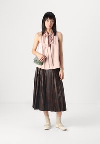 Blusa di satin rosa chiaro con dettaglio a fiocco, gonna marrone plissettata e ballerine beige. Tiene una piccola borsa a tracolla verde e nera a fantasia.