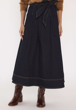 Donna che indossa una gonna midi in denim scuro con cintura annodata in vita e stivali in pelle marrone con tacco su uno sfondo neutro.
