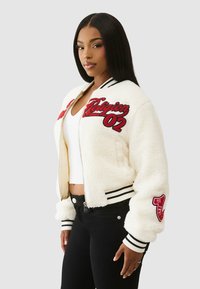 Biała kurtka bomber z polaru z czarnymi i czerwonymi haftowanymi akcentami. Ściągacze na mankietach i dole, zapinana na suwak z przodu, z charakterystycznymi naszywkami w stylu varsity.