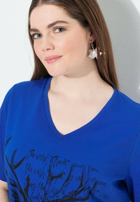 Královsky modré tričko s výstřihem do V a černými grafikami parohů a textovým potiskem na přední straně. Modelka má bílé květinové náušnice ve tvaru kruhu. Hladká textura.