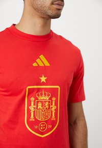 Άνδρας που φοράει κόκκινη μπλούζα με κίτρινο λογότυπο Adidas και το σήμα της ισπανικής ποδοσφαιρικής ομοσπονδίας που περιλαμβάνει στέμμα, αστέρι και ηρωικό θυρεό.