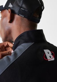Black collared shirt with gray accents, featuring a red-and-white logo on the back. Focus is on texture and attachment to a cap.