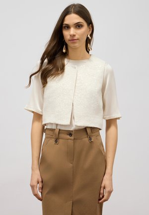 Gilet cut-off di colore avorio con un'esterno testurizzato e dettagli di paillettes. Indossato sopra una camicia chiara e abbinato a pantaloni beige.
