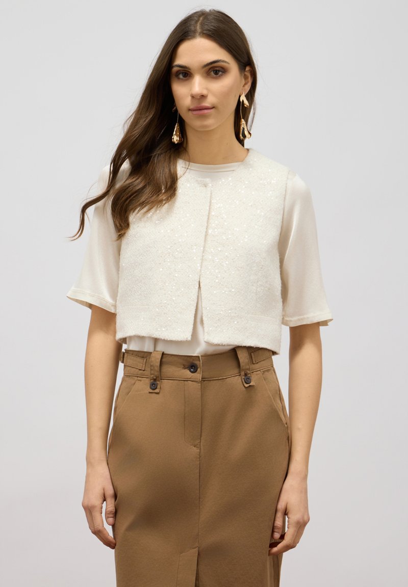 Gilet court de couleur blanc cassé avec un extérieur texturé et des accents de sequins. Porté sur une blouse clair et associé à un pantalon beige.