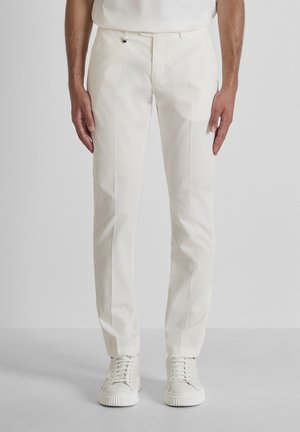 Homme portant un pantalon blanc ajusté et des baskets blanches à lacets, debout devant un fond gris uni.