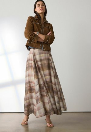 Woman standing with arms crossed wearing brown studded jacket, long plaid skirt, and beige heeled sandals against plain background.