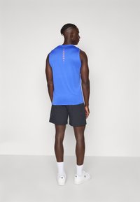 Mand står med ryggen til iført en blå ærmeløs top, sorte shorts, hvide sokker og hvide sneakers foran en ensfarvet grå baggrund.