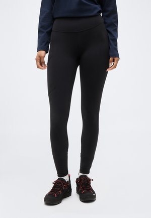 Persoană îmbrăcată cu colanți negri mulați, cu buzunar lateral, bluză cu mânecă lungă bleumarin, șosete albe și încălțăminte de drumeție neagră cu șireturi roșii.