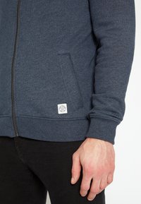 Homme portant un sweat à capuche bleu zippé avec poche avant et petit patch logo blanc, assorti à un pantalon noir.