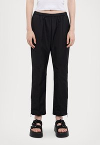 Schwarze, elastische Hose mit lockerer Passform, ausgestattet mit Reißverschlüssen an den Knöcheln und einer glatten, sleek Textur. Kombiniert mit klobigen schwarzen Sandalen.