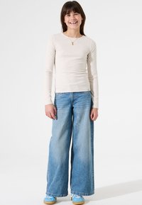 Lange mouwen beige geribbelde top gecombineerd met lichtblauwe wijde jeans. Blauwe sneakers met witte accenten ronden de outfit af. Eenvoudig ontwerp.