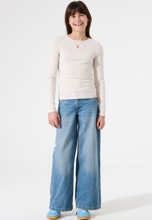 Lange mouwen beige geribbelde top gecombineerd met lichtblauwe wijde jeans. Blauwe sneakers met witte accenten ronden de outfit af. Eenvoudig ontwerp.