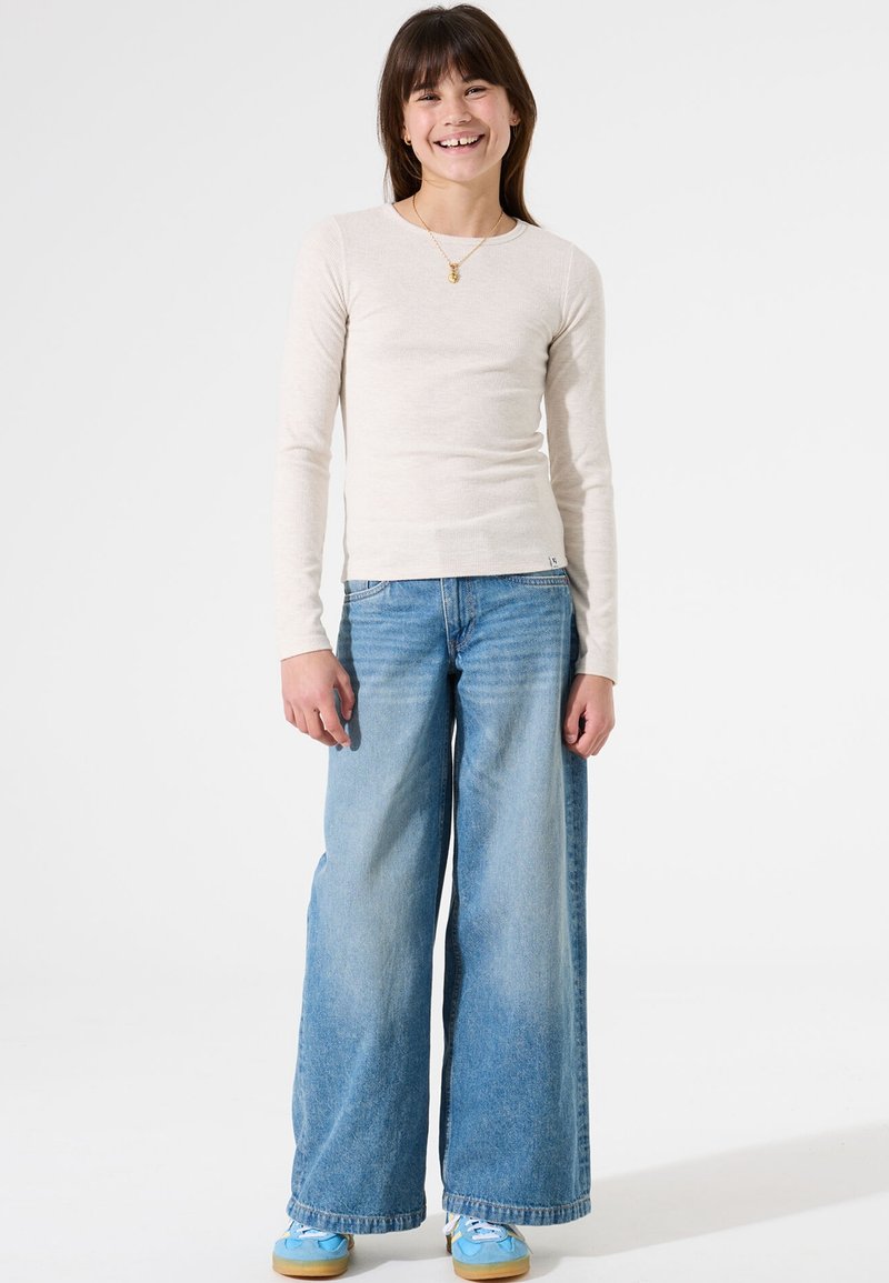 Lange mouwen beige geribbelde top gecombineerd met lichtblauwe wijde jeans. Blauwe sneakers met witte accenten ronden de outfit af. Eenvoudig ontwerp.